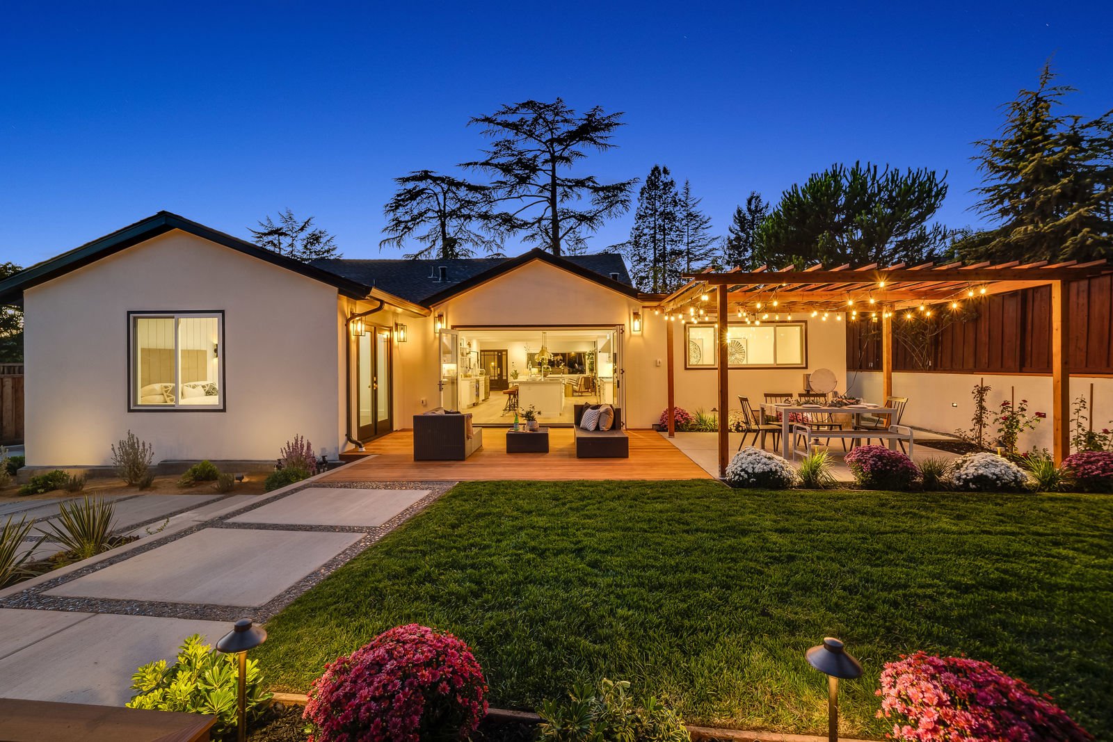 Backyard at night with pergola-covered dining area with string lights seating area on deck with opening to interior kitchen