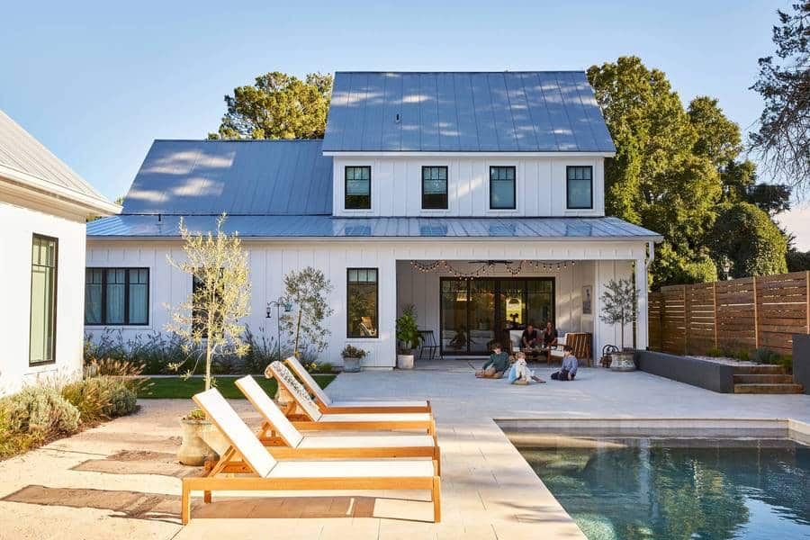 View of the backyard of a modern farmhouse style home with a with pool deck, chaise lounges and family playing in the background.